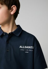 Mornarska polo majica od pamuka s kragnom, zatvaranjem na dugmad i bijelim logotipom s dodatnim jezičnim detaljima.