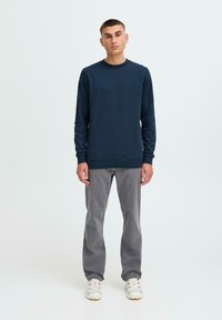 Maglione a girocollo navy con maniche lunghe, tessuto testurizzato e polsini a costine, abbinato a pantaloni grigi a gamba dritta e sneakers bianche.