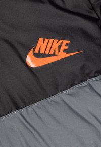 Nikejacka med svart överdel och grå underdel, med en orange logotyp på framsidan. Slät, lätt tyg med glans.