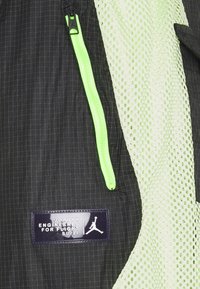 Svart jacka med grön dragkedjetrim, mesh-detaljer och en texturerad logotyp med texten "Jordan Engineered for Flight SU/21." Rutmönster synligt.