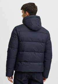 Marineblå pufferjakke med hætte, med horisontalt quiltet design, blød tekstur og elastiske manchetter. Bagfra vises sømdetaljer og silhuet.