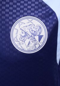 Εικονογράφηση προφίλ αρχαίου πολεμιστή που φοράει κορινθιακό κράνος μέσα σε κυκλικό πλαίσιο, με τη λέξη «AJAX» από πάνω.