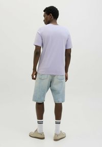 Camiseta de manga corta en color lila, de corte relajado, combinada con pantalones cortos de mezclilla clara. Calcetines blancos con rayas negras y zapatos beige completan el look.