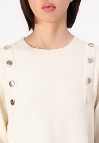 Cream knit sweater featuring a ribbed neckline and silver-tone button accents along the shoulders. Soft texture and relaxed fit.