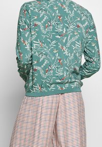 Sweat-shirt vert à manches longues avec impression florale et oiseaux, doté de poignets et d'un ourlet côtelés. À assortir avec un pantalon large à carreaux dans des couleurs pastel.