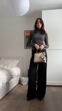 Jonge vrouw in grijze coltrui en brede zwarte broek, met een beige handtas met sjaal, in een minimalistische slaapkamer met een wit bed en een poster.