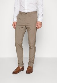 Beige tailored trousers with a smooth texture, featuring a button closure and front pockets, paired with brown loafers and a white patterned shirt.