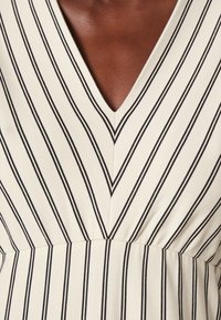 Krémové šaty s výstřihem do V, které mají vertikální a diagonální tenké černé pruhy a shromážděný pas na horní části těla tmavé pleti.