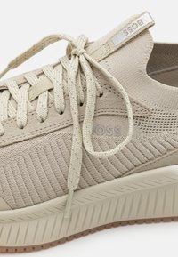 A bézs sneaker textil felsőrésszel rendelkezik, bordázott mintázattal, lapos fűzőkkel és barázdált talppal. Az oldalán található márkajelzés jól látható.