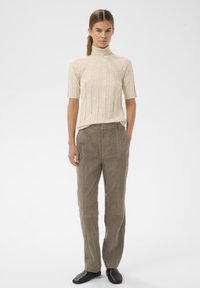 Vrouw staand, gekleed in een beige korte mouwen coltrui, taupe broek en zwarte instapschoenen, tegen een effen witte achtergrond.