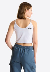 Femme aux cheveux châtain clair porte un débardeur court blanc et un pantalon bleu à taille élastique, vue de dos montrant le dos et le profil de côté.