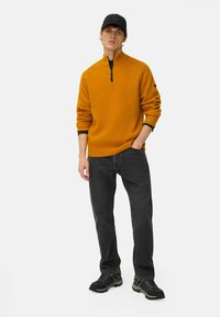 Oranger ribbelter Zip-Hals-Pullover, schwarze Jeans und schwarze Sneakers. Der Pullover hat einen hohen Kragen und eine strukturierte Oberfläche; die Jeans haben eine klassische Passform.