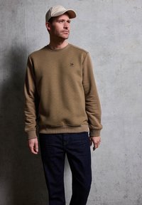 Felpa marrone con polsini e orlo a costine, con un piccolo logo. Jeans blu scuro e un cappellino beige chiaro completano l'outfit.