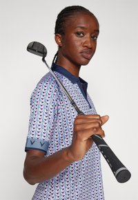 Patterned golf shirt in blue tones with geometric design, paired with a golf club. Features a navy collar and textured black grip.