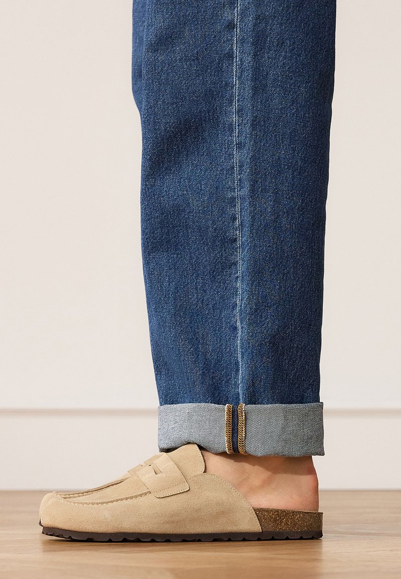 Personne portant un jean bleu retroussé et des mocassins beiges en suède sans lacets, debout sur un sol en bois contre un mur uni.