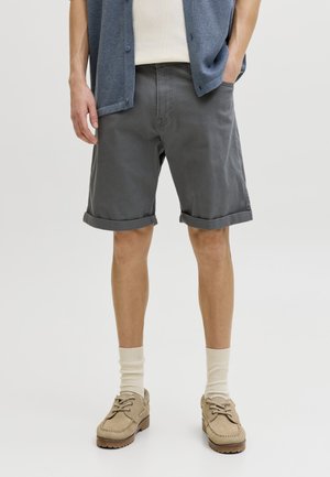 Personne portant un short gris retroussé, des chaussettes crème, des mocassins beiges, une chemise blanche et une chemise bleue ouverte. Mains dans les poches.