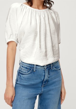 Femme portant un chemisier blanc avec un col froncé et de courtes manches bouffantes, associé à un jean en denim bleu taille haute.