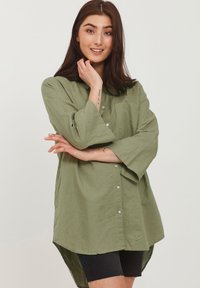 Camicia in lino verde oliva con colletto, maniche lunghe e bottoni argentati, con taschino frontale e orlo curvo.