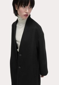 Abrigo de lana negro con una silueta relajada, que presenta un escote en V profundo, botones en la parte frontal y bolsillos laterales. Combinado con un cuello de tortuga blanco.