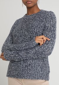 Gestrickter Pullover mit Rundhalsausschnitt, gefertigt aus dickem, strukturiertem Garn in Blautönen. Verfügt über gerippte Bündchen und Saum sowie seitliche Schlitze am unteren Rand.