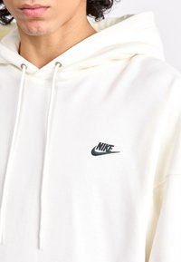 Baltas Nike megztinis su minkšta tekstūra, su juodu siuvinėtu logotipu krūtinėje ir kapišono raišteliais.