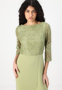 Robe verte avec un corsage en dentelle à motifs floraux et des manches trois-quarts, se dégradant en une jupe verte unie. Texture lisse.