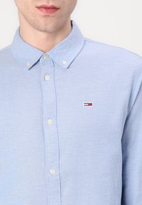 Ljust blå skjorta med knapp-up, krage, vita knappar och en liten broderad logotyp med röd, vit och blå detaljer.