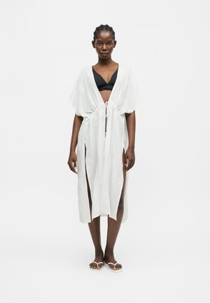 Mujer con bikini negro bajo un protector blanco hasta la rodilla, abierto por delante, con cinturón atado a la cintura y aberturas laterales, combinado con chanclas blancas.