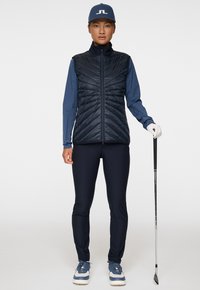 Takkahuppu sininen pitkähihaisessa paidassa, yhdistettynä mustiin housuihin ja vaaleansinisiin lenkkareihin. Golfmaila kädessä, päässä lippis.