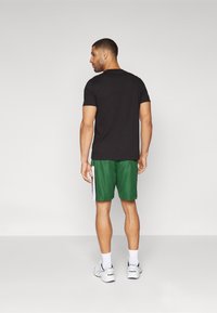Chemise noire à manches courtes, short de sport vert avec des bandes blanches sur les côtés, et sneakers blanches. Sujet tourné dos à la caméra.