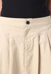 Beige plissert skjørt med flat midje, med svart knappelukking. Materialet ser lett og glatt ut, med sidelommer.