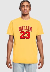 Žuta pamučna majica s velikim crvenim tekstom koji glasi "BALLIN" i brojem "23." Klasični okrugli izrez i kratki rukavi.