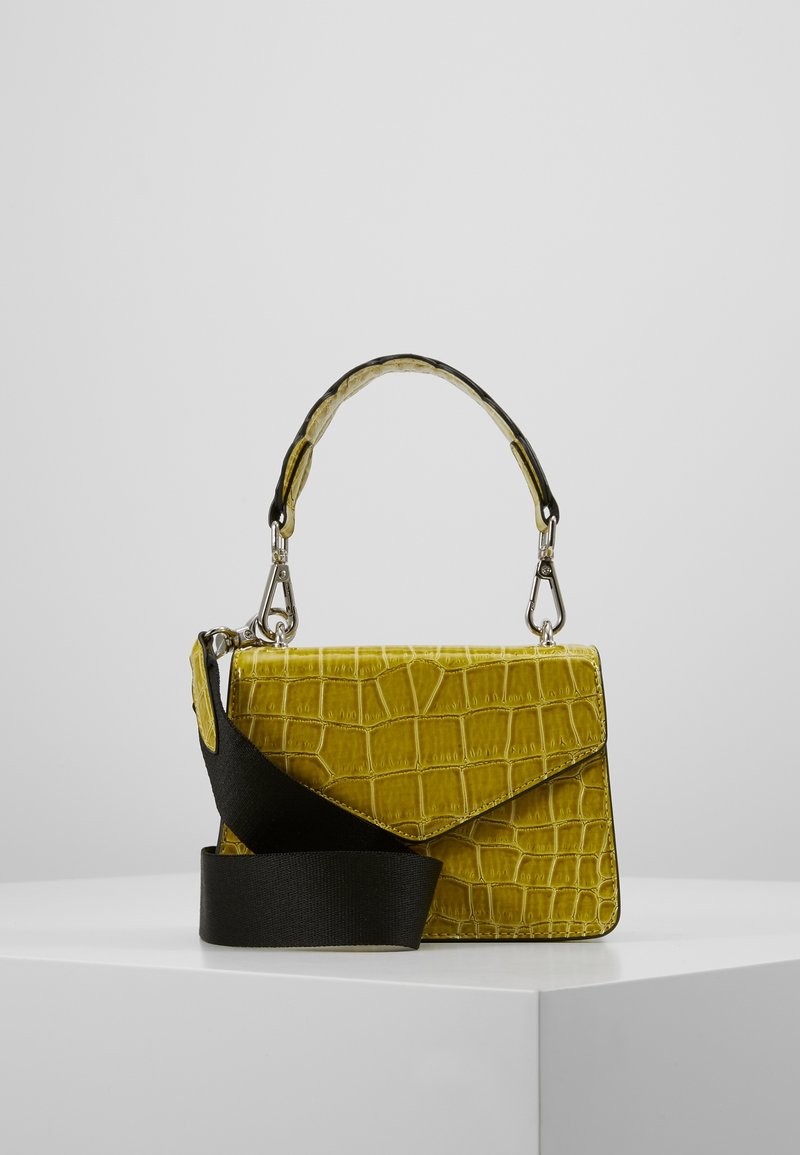Sac à main jaune à texture crocodile avec poignée supérieure et large bandoulière noire, présenté sur un socle blanc sur fond uni.