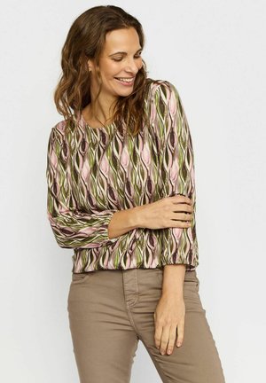 Smilende kvinne med bølgete brunt hår som har på seg en mønstret langermet topp i rosa, grønt og brunt, kombinert med beige bukser, står foran hvit bakgrunn.