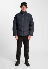 Granatowa kurtka puchowa z wysokim kołnierzem, pikowanym wzorem i pełnym suwakiem. Połączona z czarnymi spodniami i czarną dzianinową czapką.