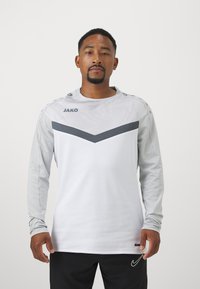 Langarm sportshirt in het wit met een grijs chevronontwerp. Gemaakt van ademend materiaal, met een logo op de borst en schouderknopen.