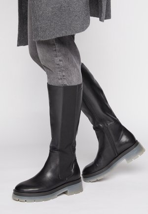 Personne portant des bottes en cuir noires hautes jusqu’au genou avec des semelles épaisses grises, associées à un jean gris et un manteau gris.