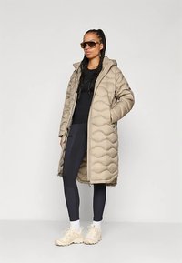 Beige kapucnis steppelt téli kabát oldalzsebekkel. Fekete felső és leggings fölött viselve, világos sportcipővel és napszemüveggel párosítva.