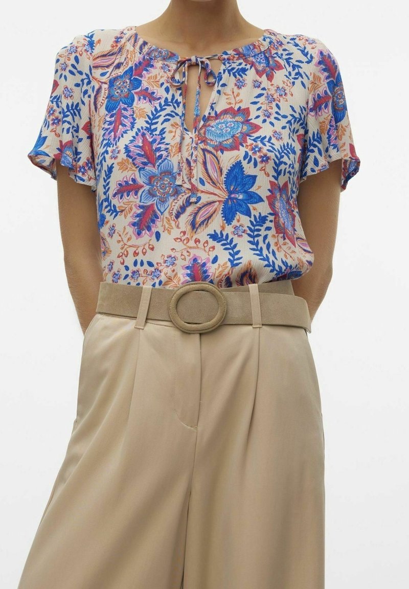 Blumenbluse in Creme mit blauen und korallenfarbenen Mustern, mit einer gebundenen Halslinie. Kombiniert mit beigen hochgeschnittenen Hose und einem runden Gürtel.