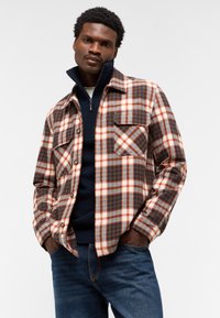 Karierte Flanellbluse in Braun, Orange und Marineblau, mit zwei Brusttaschen und Knopfleiste, über einem Marineblauen Pullover getragen.