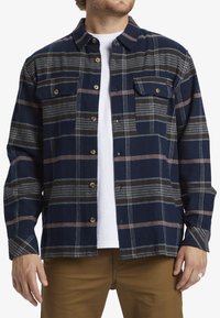 Camisa de franela azul marino con un patrón de cuadros y rayas blancas y naranjas, dos bolsillos en el pecho y cierre de botones. Material de algodón, corte relajado.