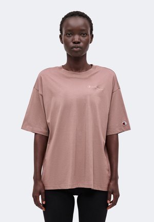 Kvinde med kort hår iført en løs lysebrun Champion t-shirt og sorte bukser, der står foran en ensfarvet lys baggrund.