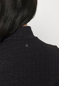 Gros plan sur un haut en tissu côtelé foncé avec des paillettes subtiles porté par une personne aux longs cheveux foncés, montrant un petit logo métallique "G" près du col.