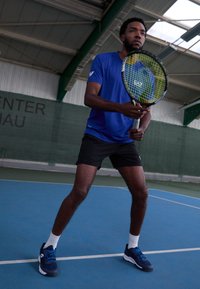 Camisa deportiva azul, pantalones cortos negros, calcetines blancos y zapatillas de tenis azules. Sosteniendo una raqueta de tenis con un patrón de cuerdas verde y negro. Pista cubierta.