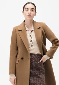 Bruin wollen jas met een gelapte kraag, dubbele knopen en zijzakken, gelaagd over een beige overhemd en een gestructureerde, gemusterde rok.