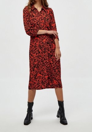 Femme portant une robe midi fleurie rouge et noire avec des manches trois-quarts et des bottines noires, debout devant un fond uni.