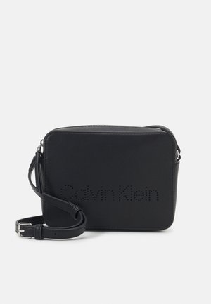 Kompaktowa czarna prostokątna torba na ramię z regulowanym paskiem i perforowanym logo Calvin Klein z przodu.