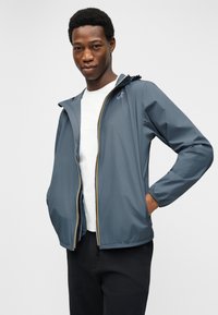 Νεαρός άνδρας που φοράει μπλε ζακέτα με κουκούλα πάνω από λευκό μπλουζάκι και μαύρο παντελόνι, με τα χέρια στις τσέπες της ζακέτας, μπροστά σε λευκό φόντο.