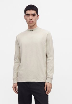 Junger Mann trägt ein hellbeiges Langarmhemd mit dem Text "BOSS" am Kragen und schwarze Hose vor einem schlichten weißen Hintergrund.