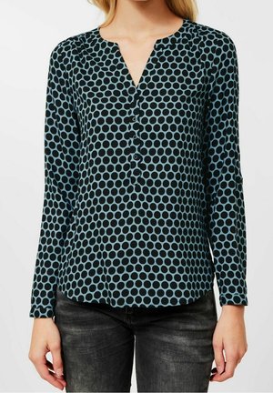 Frau trägt eine langärmelige blaue Bluse mit schwarzen Pünktchen und schwarze Jeans, steht vor einem einfarbigen weißen Hintergrund.